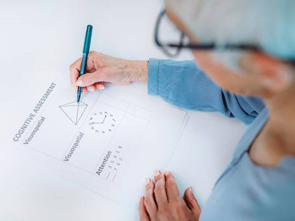 Elderly lady drawing a shape on a sheet of paper entitled Cognitive Assessment