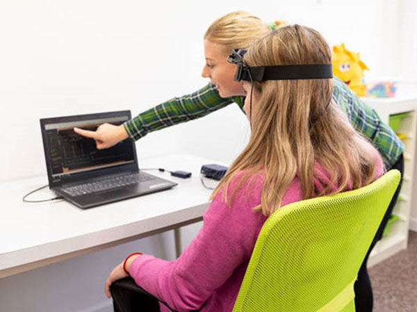 A researcher is pointing to a computer screen. A teenage girl is sitting in front of the computer wearing a head device.