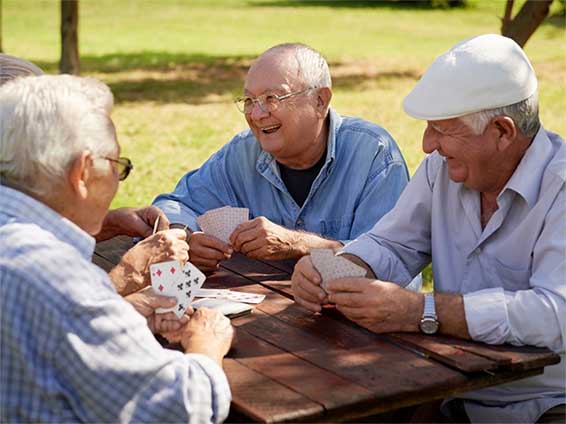 Three elderly men are sitting at a table in a park. They are playing cards and laughing.